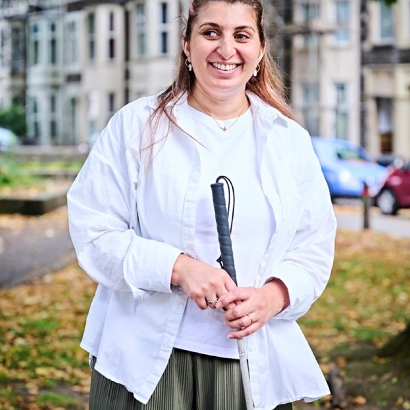 Tasfsila is a South Asian female in her 40s is standing smiling in a park area holding her long white cane. She’s wearing a white t-shirt with a white open shirt and khaki green trousers.