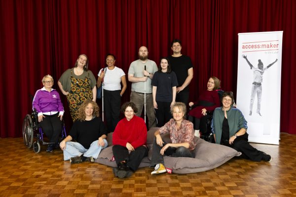 11 workshop presenters and team members pose for a photo next to an access maker banner. The diverse group are made of up wheelchair users and visually impaired people with various people standing and sitting on bean bags for the photo.