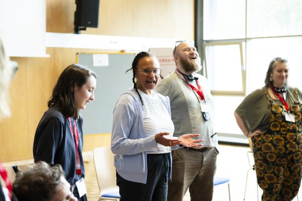 Maria talking in a workshop space to participants, with Ellen and Ben standing and laughing next to her.