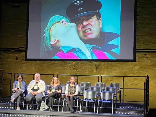 Maria sits on the stage next to three panel members. Behind her is a large project screen displaying an image from Extant’s production ‘Resistance’ which displays a close up image of a man grimacing in a police man’s hat, pulling back the head of another man, wearing a flat cap.