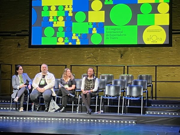 Maria sits on the stage next to three panel members. Behind her is a large project screen displaying the festivals logo with a variety of blue, green and yellow squares.