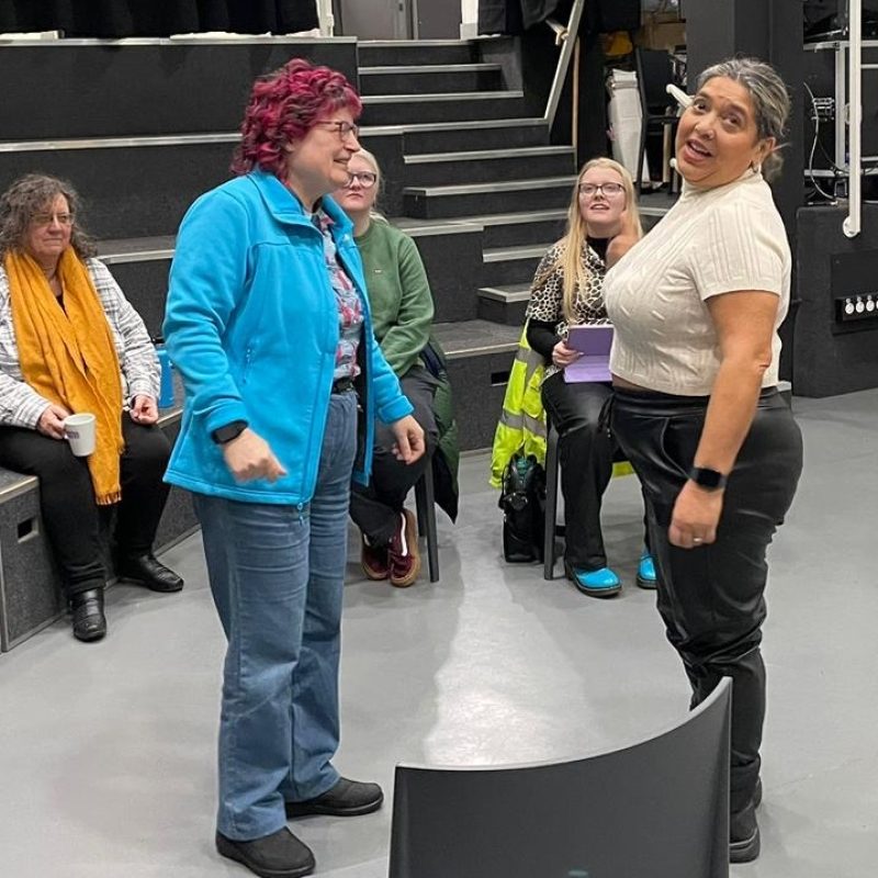 Two women are acting mid-scene, behind them three audience members are watching on intently.