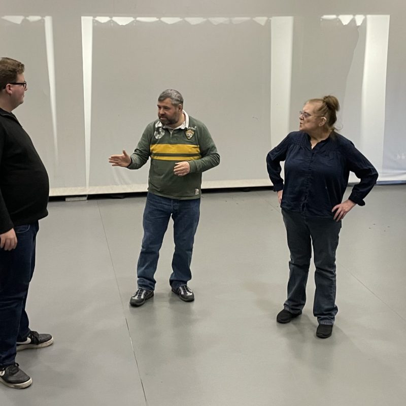 Three people are standing in the space about to perform to an audience. The facilitator stands in between them explaining the task.