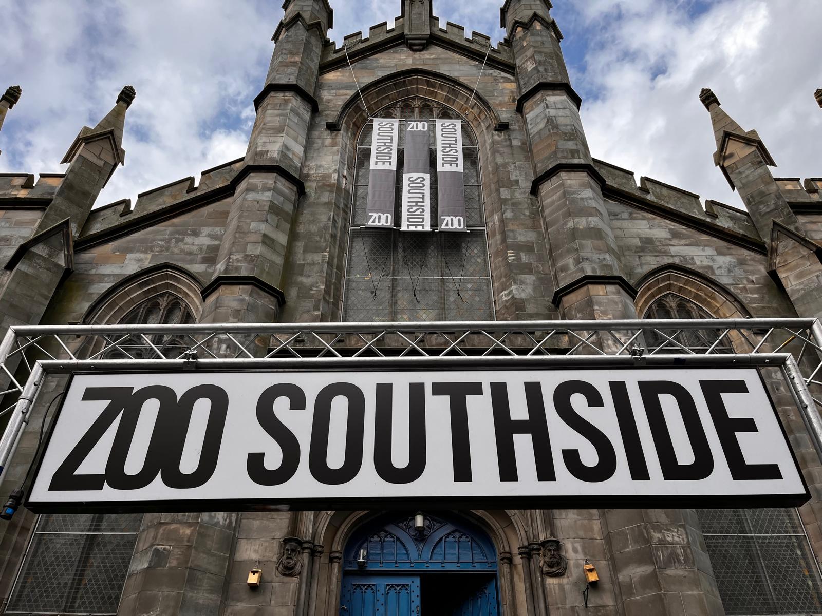 Exterior of ZOO Southside, a large stone church-style building with pointed towers and arched windows. A sign reading 'ZOO SOUTHSIDE'in bold black letters on a white background hangs above the entrance.
