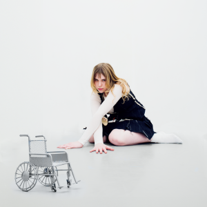 Against a white background, a white woman with brown hair and blonde highlighted streaks kneels on the floor, with her arms outstretched and her palms touching the white floor. She wears a long sleeved white t-shirt and a black jumpsuit. At the bottom left is a silver wheelchair.