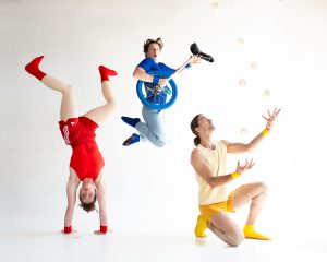 3 people are performing circus tricks. To the left a person is wearing all red and is performing a handstand, in the middle, a person is wearing all blue, is jumping in the air, holding a tri-cycle and to the right, a person is wearing yellow and kneels with holding their arms up in the air. 