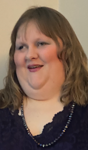 Ava is a young white woman. She has long curled light brown hair, wears a necklace and a navy lace-patterned v shaped dress.