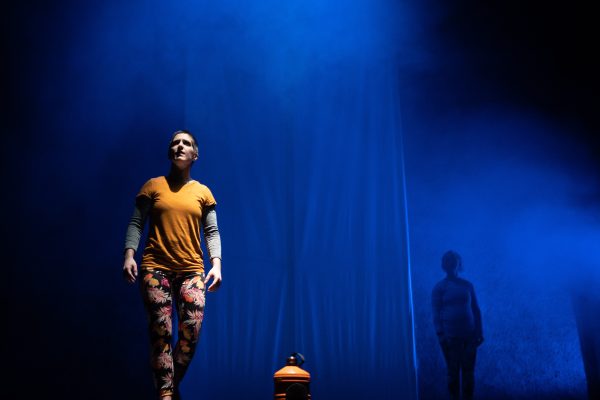 A wide landscape shot of two white people onstage. One is fully lit and wearing yellow. The other in shadow. The stage is lit a smokey blue.