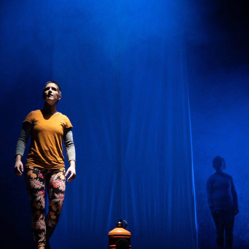 A wide landscape shot of two white people onstage. One is fully lit and wearing yellow. The other in shadow. The stage is lit a smokey blue.