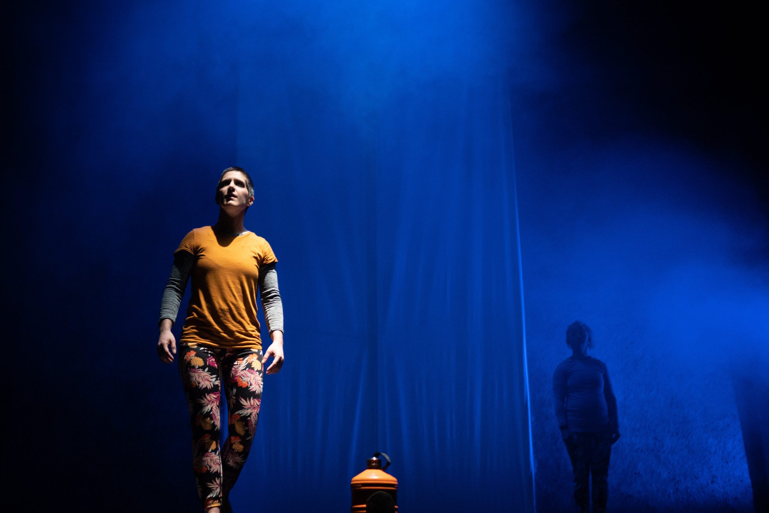 A white person stands onstage, fully lit in the spotlight and wearing yellow. The stage is lit a smokey blue and on the floor an orange water cooler bottle.