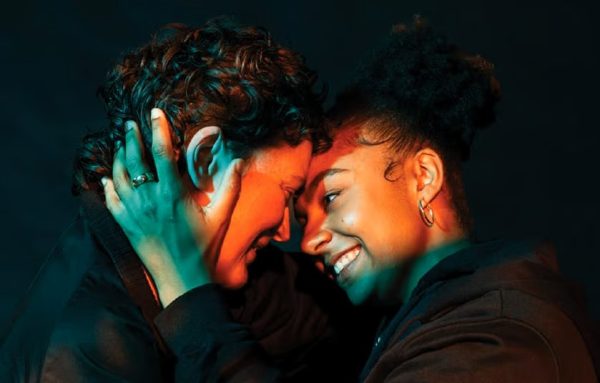 A close up of two people’s faces as they smile at each other, shot in all black environment. The person on the right is cradling the other persons face. Their faces are illuminated by an orange spotlight.