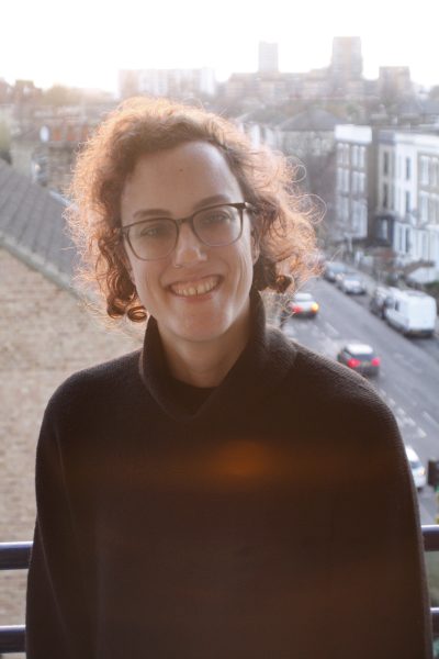 A person with short curly brown hair, glasses, and a nose ring is smiling warmly at the camera. They are wearing a dark sweater and standing on a balcony overlooking a London street lined with terraced houses. The background is softly lit by the setting sun, casting a golden glow over the scene. 