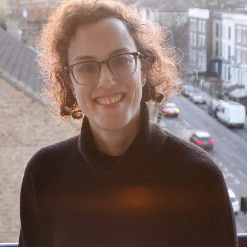 A person with short curly brown hair, glasses, and a nose ring is smiling warmly at the camera. They are wearing a dark sweater and standing on a balcony overlooking a London street lined with terraced houses. The background is softly lit by the setting sun, casting a golden glow over the scene.