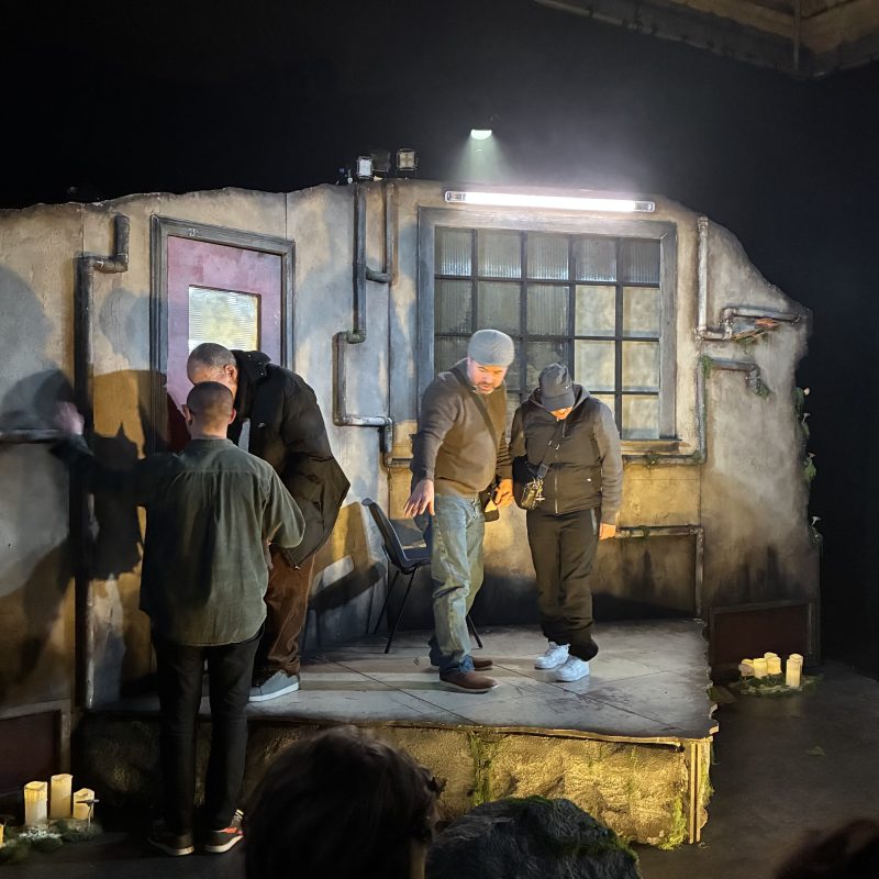 During the touch tour, 4 people are standing on the set of ghost stories, which is a dark grimy room with pipes on the wall and a raised floor. To the right a large window with smaller square panels and to the left a red door. Around the set are lit up candles.