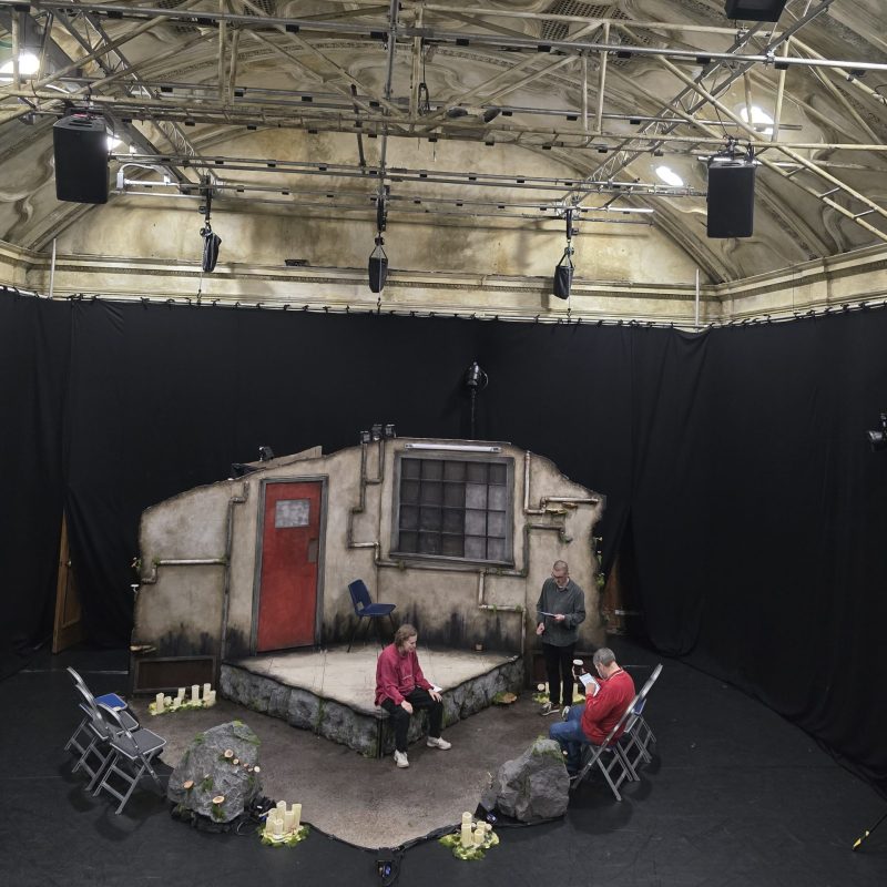 A wide angle shot of the stage space. A simple set showing a grimy room with pipes on the wall and a dirty mattress on the floor is situated at the centre of an all black stage. One person sits in the mattress, one stands near them and one sits on a chair in front of the stage. Above them the set are the arched white walls of the BAC theatre space with a lighting rig.