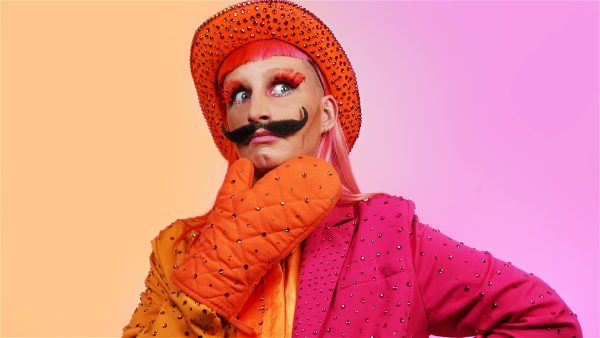 A person wearing a brightly coloured bejeweled half orange and half pink outfit. They wear an organe bejeweled oven glove and hold their hand up to their chin. They wear an orange bejeweled cowboy hat. They have a black curly moustache.