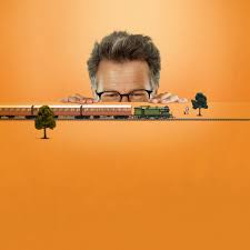 On an orange background, a white person with short hair and glasses is peering at a railway track with a train model and small trees.