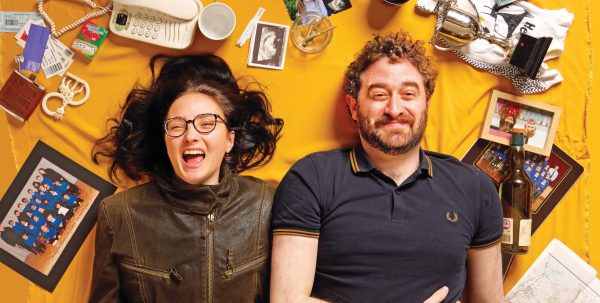 Theatre image for Scenes from a Friendship. A man and woman are lying on a yellow sheet with wide facial expressions and household objects, photos and memorabilia surrounding them.