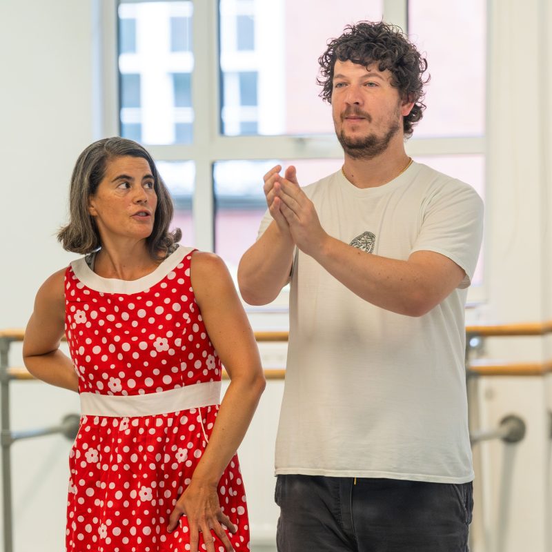 Two people, Eva and Alberto, stand in a dance studio. One wears a red polka-dot dress with a white belt, the other a white T-shirt and dark pants, with hands raised as if gesturing.