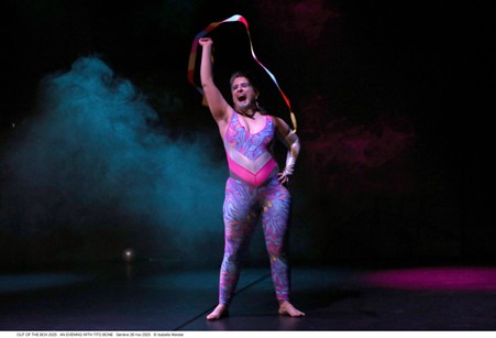 Tito Bone, an average build white drag king with a brown and grey mullet and teal glitter beard stands with a big openmouthed smile twirling a colourful ribbon above their head. They are barefoot and wear a pink and blue cat suit.