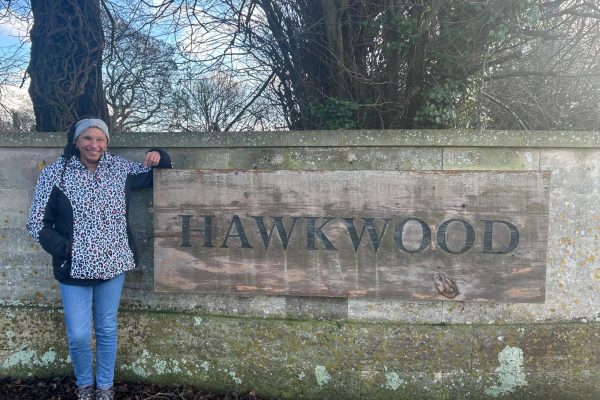 Maria is leaning and smiling next to a large Hawkwood wooden sign.