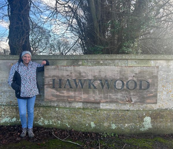 Maria is leaning and smiling next to a large Hawkwood wooden sign.