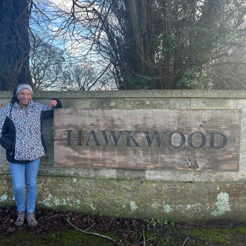 Maria is leaning and smiling next to a large Hawkwood wooden sign.