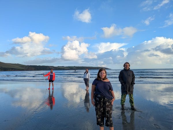 Extant team on Par Beach, from left to right Maria, Louisa, Tam and Ian. They face towards the camera, Maria holds her white cane. They stand in the water, with the waves behind them. In the backdrop, a blue sky with white fluffy clouds.