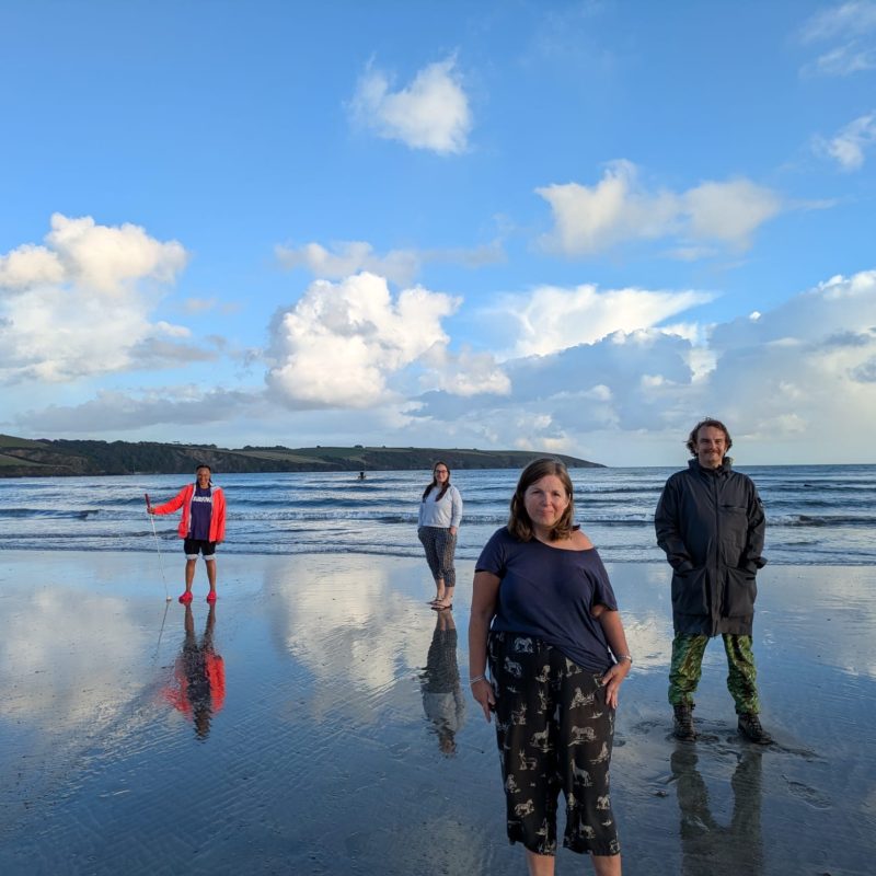 Extant team on Par Beach, from left to right Maria, Louisa, Tam and Ian. They face towards the camera, Maria holds her white cane. They stand in the water, with the waves behind them. In the backdrop, a blue sky with white fluffy clouds.