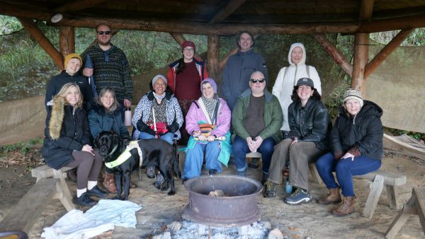 Sitting around a firepit, a group of 12 people are smiling and posing for the photo. They are wearing thick jumpers, coats and woolly hats. The group consists of collective dreamers, Extant staff and access workers. A black guide dog is present in the picture.