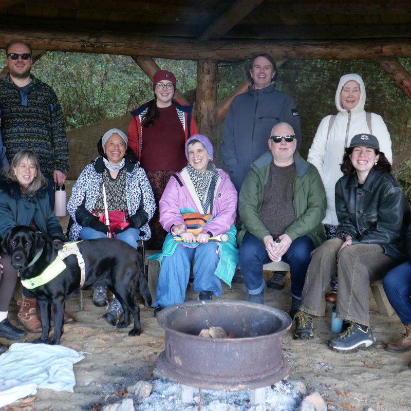 Sitting around a firepit, a group of 12 people are smiling and posing for the photo. They are wearing thick jumpers, coats and woolly hats. The group consists of collective dreamers, Extant staff and access workers. A black guide dog is present in the picture.