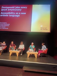 Maria talking on the panel on stage, with the panel members listening. On the Screen 'Accessibility as a new artistic language' is displayed.