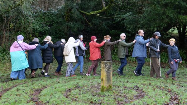 A group of 12 people, wrapped up in warm clothing are outside in a long line, holding their right arm on the person in front’s right shoulder.