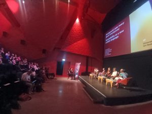 A side on wide shot of Maria talking to the audience and other panellists in view.