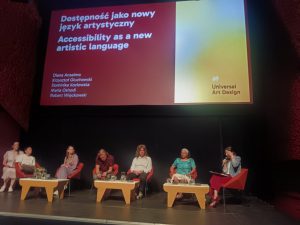 Maria is sitting on stage with the conference panel members. On the screen, 'accessibility as a new artistic language' is displayed.