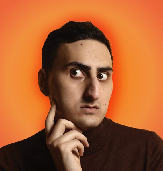 Aarian Mehrabani’s face against an orange background. They are wearing a black shirt and hold their finger up to their cheek.