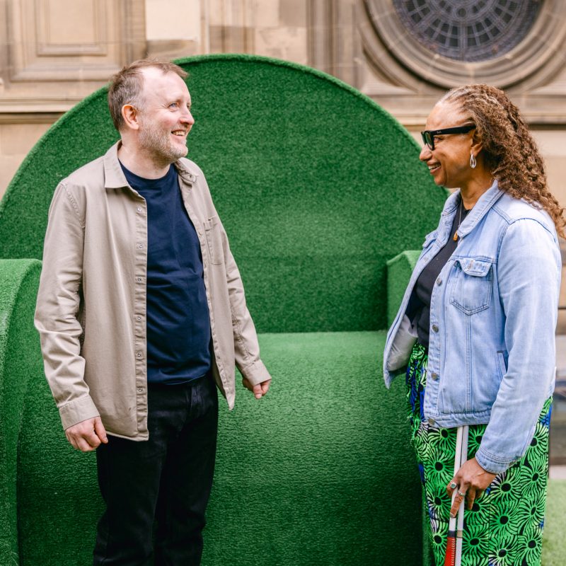 Chris McCausland and Dr Maria Oshodi standing together in discussion. Chris is a white man wearing an open long sleeve beige shirt, a navy-blue shirt and black trousers. Maria is a mixed heritage brown skin woman, wearing a navy t-shirt and blue and green peacock patterned skirt and she holds her cane. Behind them is a giant green chair.