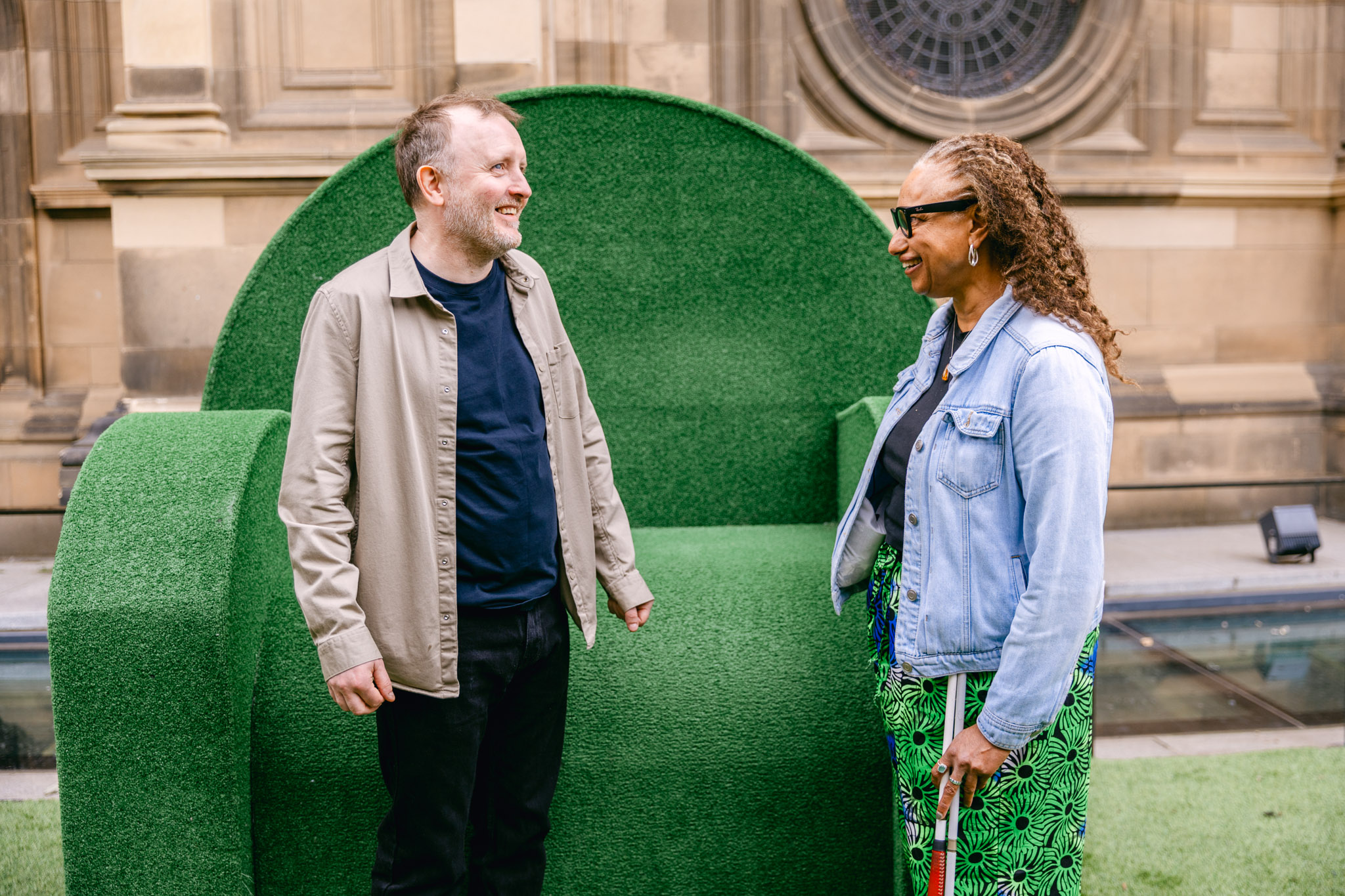 Chris McCausland and Dr Maria Oshodi standing together in discussion. Chris is a white man wearing an open long sleeve beige shirt, a navy-blue shirt and black trousers. Maria is a mixed heritage brown skin woman, wearing a navy t-shirt and blue and green peacock patterned skirt and she holds her cane. Behind them is a giant green chair.