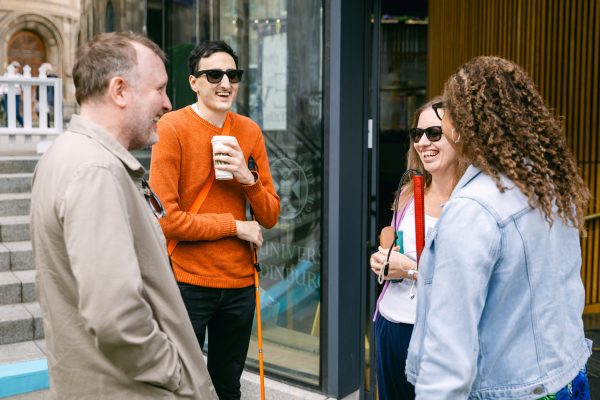 A group of four people laughing. From left to right: Chris McCausland, Aarian Mehrabani, Holly Thomas and Dr Maria Oshodi. They are wearing meta glasses and Maria and Aarian are holding their canes. 