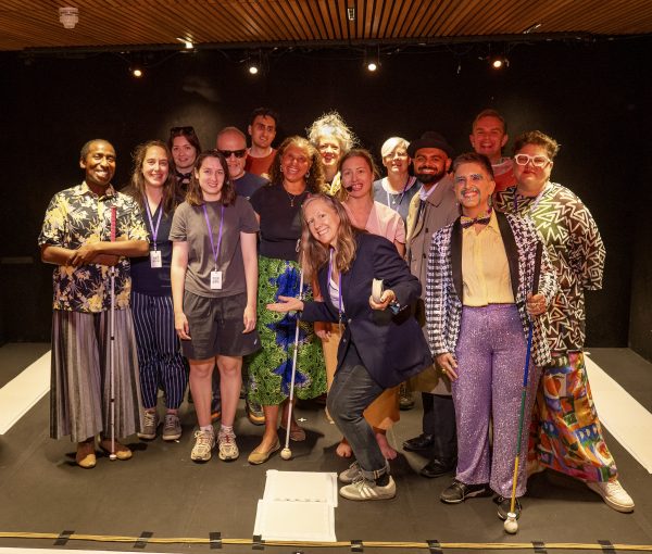 A group of 15 visually impaired artists and Extant team members are smiling for a photo on a black box theatre stage. They are a diverse group of people, and at the front three people are holding canes.
