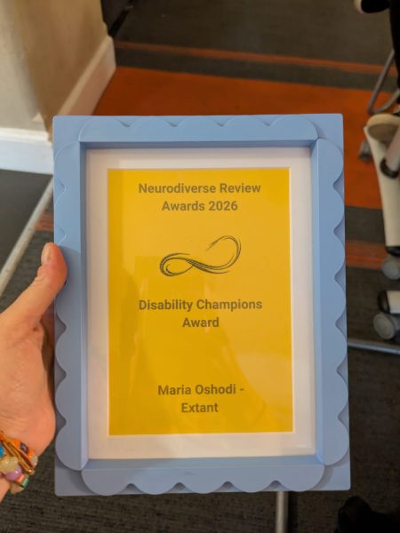 A hand holds the Disability Champions Award Framed Certificate. It is a light blue photo frame with scallop edges, and with a yellow background and black text, which reads ‘Neurodiverse Review Awards 2026’. Below this is the Neurodiverse reviews logo and then black text, which reads ‘Disability Champions Award’ and ‘Maria Oshodi – Extant’. 