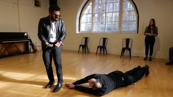 Chris lies outstretched on the floor; his arms stretch towards Shashank's feet. Shashank's is speaking to Chris and holds the palms of his hands facing forward.