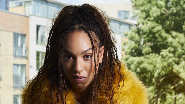 A woman stares at the camera. She wears a yellow fluffy coat. Behind her is a building and trees.