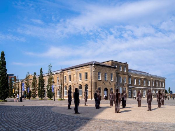 The courtyard of Woolwich Works, a cobblestone plaza with human size statues. The statues stand in front of the Georgian brick building, which has large arched windows and doorways.