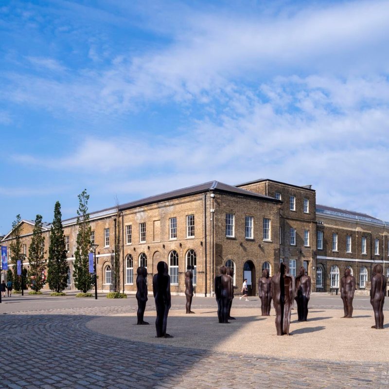 The courtyard of Woolwich Works, a cobblestone plaza with human size statues. The statues stand in front of the Georgian brick building, with large arched windows and doorways.