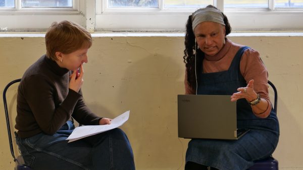Grace and Maria sitting down. Grace has her finger on cheek, and looks at Maria, with a script in her hand. Maria holds her laptop on her lap and holds out her hand.