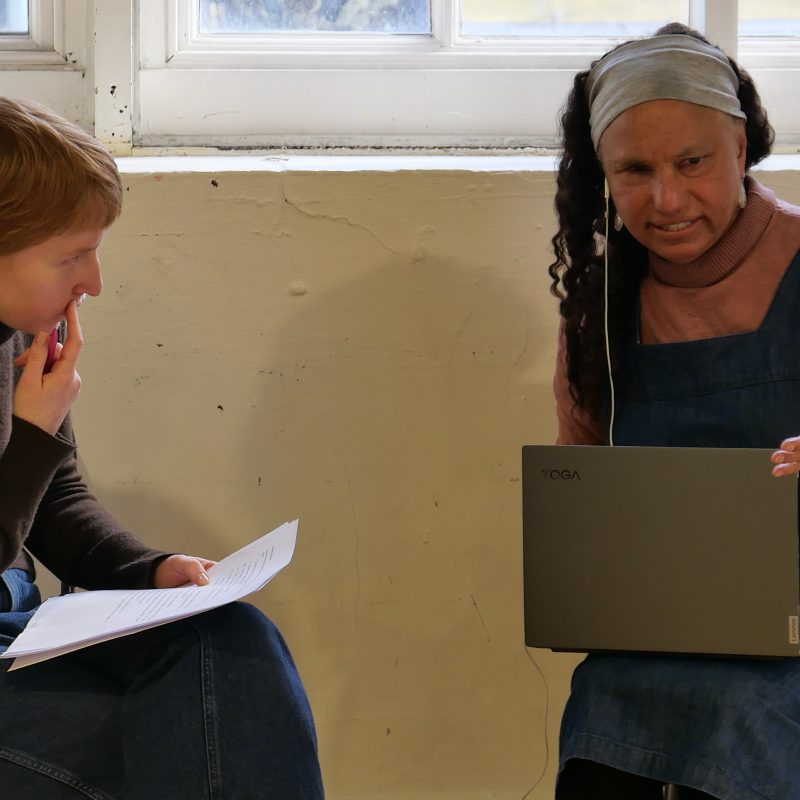 Grace and Maria sitting down. Grace has her finger on cheek, and looks at Maria, with a script in her hand. Maria holds her laptop on her lap and holds out her hand.