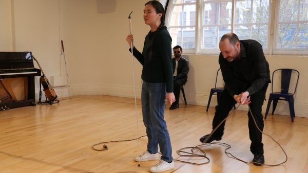 Jasmine holds her cane and is mid talking, behind her Chris is crouched, playing with a brown thin piece of rope.