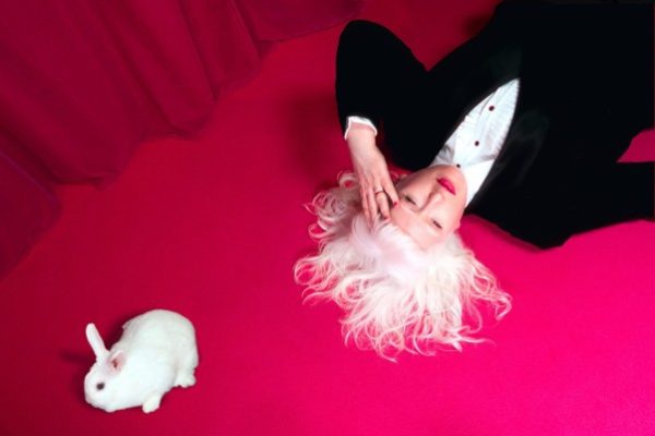 On pink flooring, a women, with long white hair, wearing black jacket and white shirt lies down. She places her hand on her head. Next to her is a white rabbit.