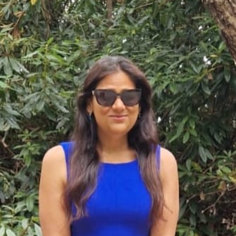 A dark-haired woman in her mid-thirties with olive toned skin and mid length wavy locks cascading down either side of her shoulders. She is wearing a sleeveless sapphire blue dress and sunglasses, affecting a soft smile while standing in front of lush greenery.